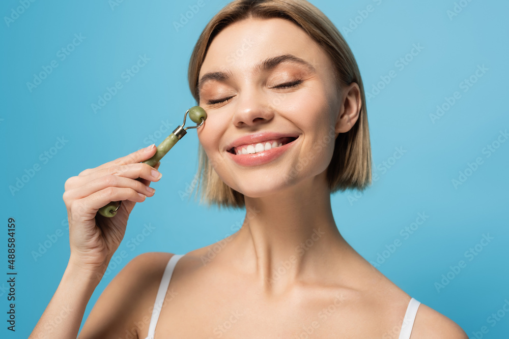cheerful young woman massaging face with jade roller isolated on blue cheerful young woman massaging face with jade roller isolated on blue