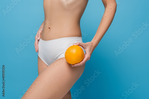 cropped view of slim young woman in white lingerie holding whole orange isolated on blue