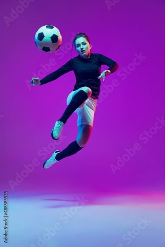 One sportive girl, female soccer player kick in jump football ball isolated on purple studio background in neon light. Sport, action, motion, fitness