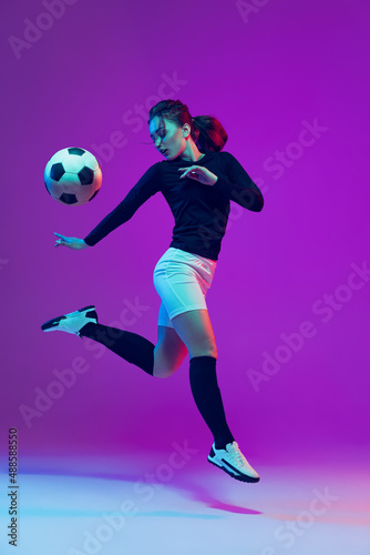 Dynamic portrait of female soccer player practicing with football ball isolated on purple studio background in neon light. Sport, action, motion, fitness