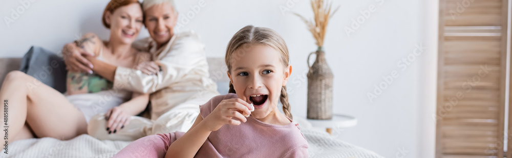 Kid holding popcorn near blurred lesbian parents on bed, banner Kid holding popcorn near blurred lesbian parents on bed, banner