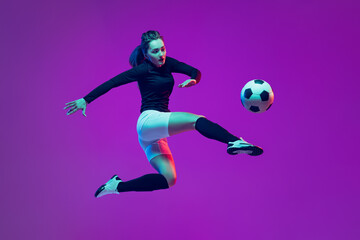 One sportive girl, female soccer player kick in jump football ball isolated on purple studio background in neon light. Sport, action, motion, fitness