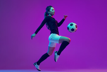 Dynamic portrait of female soccer player practicing with football ball isolated on purple studio background in neon light. Sport, action, motion, fitness