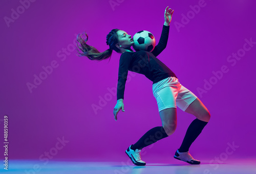 Emotive girl, female soccer, football player in motion, action isolated on purple studio background in neon light. Sport, active, healthy lifestyle