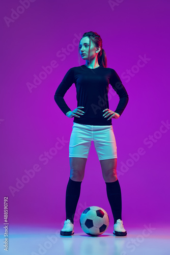 Portrait of beautiful girl, professional female soccer player posing with football ball isolated on purple studio background in neon light. Sport, team, games, fitness © master1305