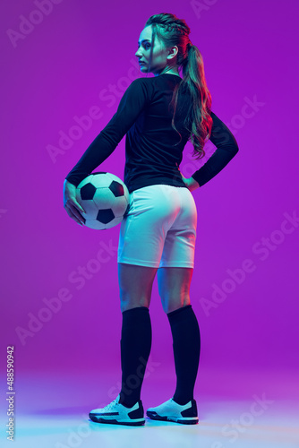 Portrait of beautiful girl, professional female soccer player posing with football ball isolated on purple studio background in neon light. Sport, team, games, fitness © master1305
