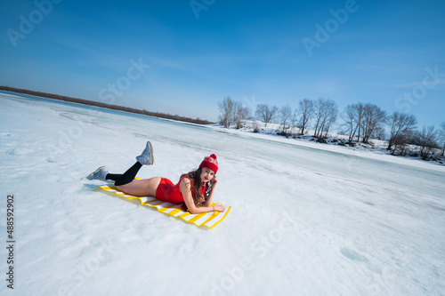 Caucasian woman in a red swimsuit and a knitted hat sunbathes in winter lying on the snow.