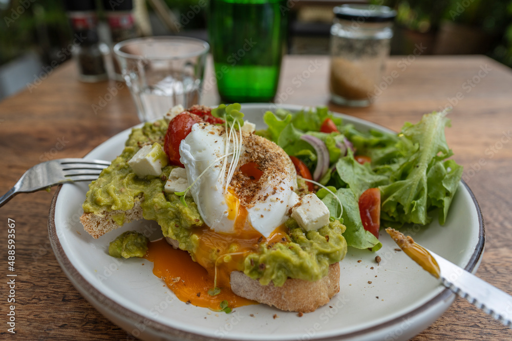 Sourdough toast, poached eggs, avocado pulp and fresh vegetables on plate in cafe, close up Sourdough toast, poached eggs, avocado pulp and fresh vegetables on plate in cafe, close up