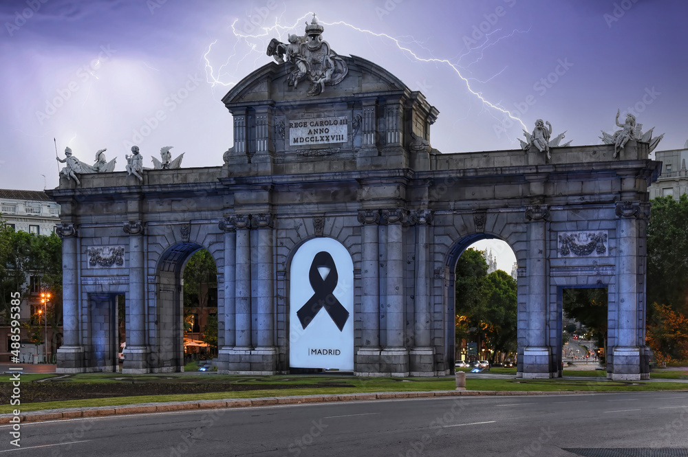 Vista de la Puerta de Alcalá en Madrid con su arco central tapado con un cartel recordando a los muertos por Covid bajo un cielo iluminado por relámpagos