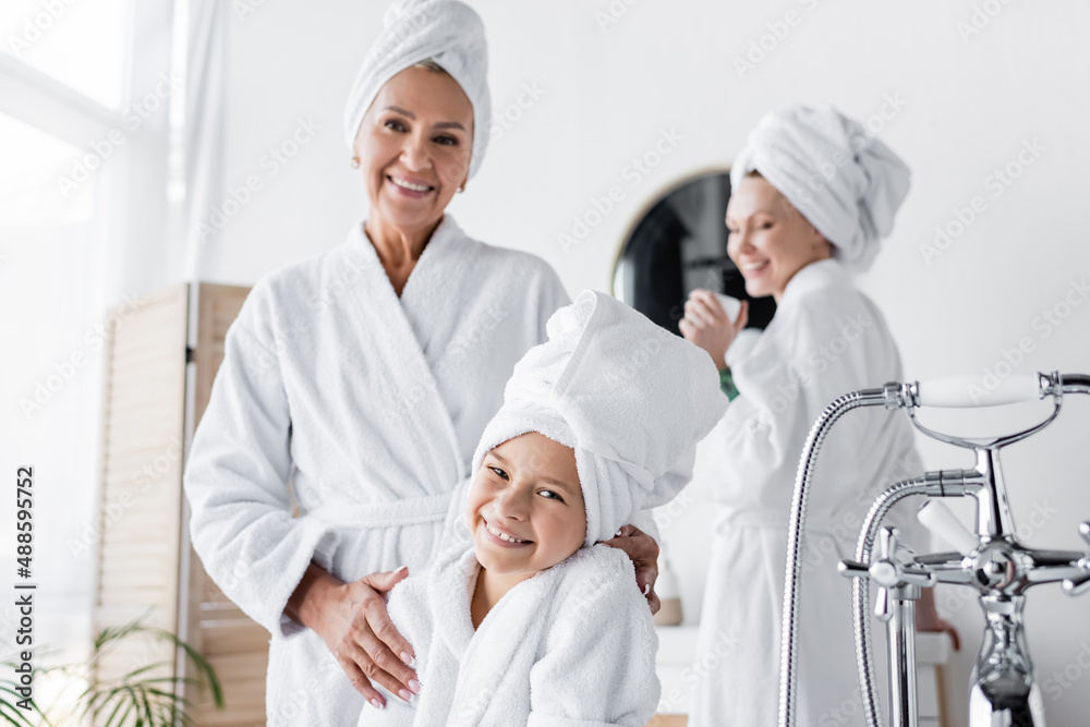 Smiling kid in towel and bathrobe looking at camera near mom at home
