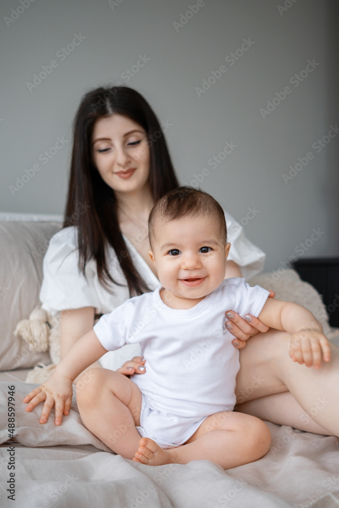A young mother plays with her little child sitting on the bed. A smiling baby is sitting on a bed with his mother in a country house. A 9-month-old girl looks into the camera. A young mother plays with her little child sitting on the bed. A smiling baby is sitting on a bed with his mother in a country house. A 9-month-old girl looks into the camera.