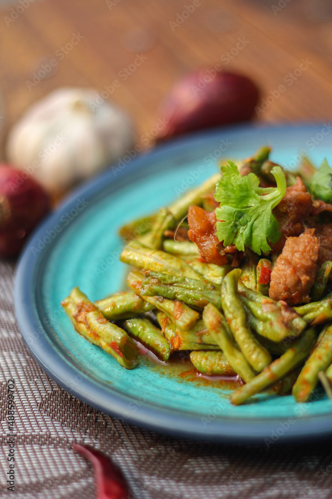 Stir fried pork belly and red curry paste with sting bean served in a blue plate on a wooden table. Stir fried pork belly and red curry paste with sting bean served in a blue plate on a wooden table.