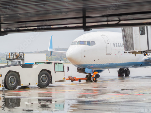 A man in uniform attaches an airfield tractor to an airliner on a rainy day. Aircraft towing