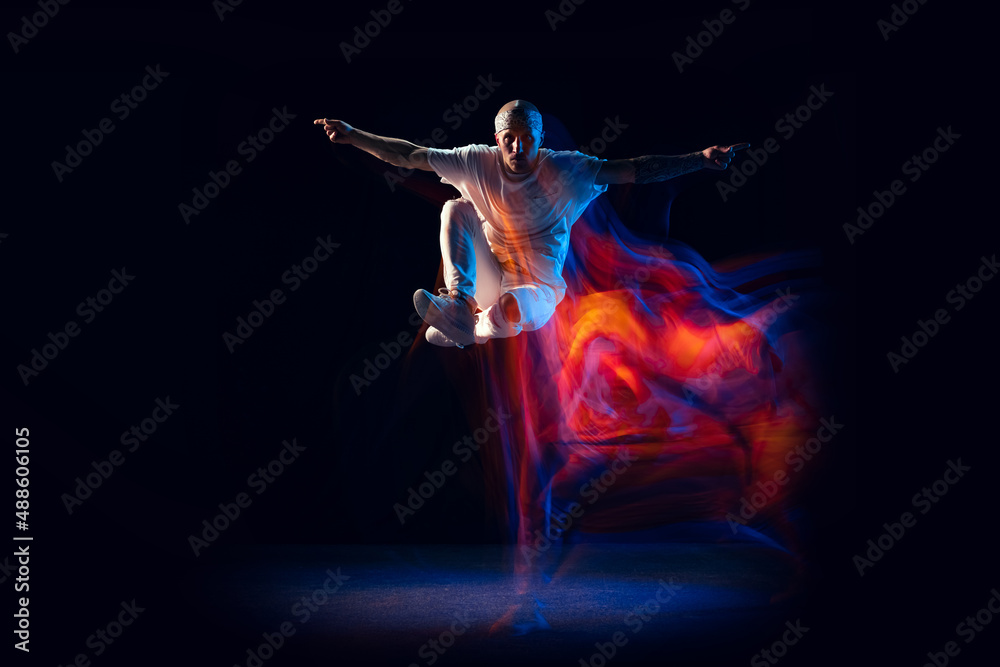 Jumping. Stylish man in sports white outfit dancing hip-hop, breakdance isolated on dark background in mixed neon light. Youth culture, hip-hop, style and fashion, action.