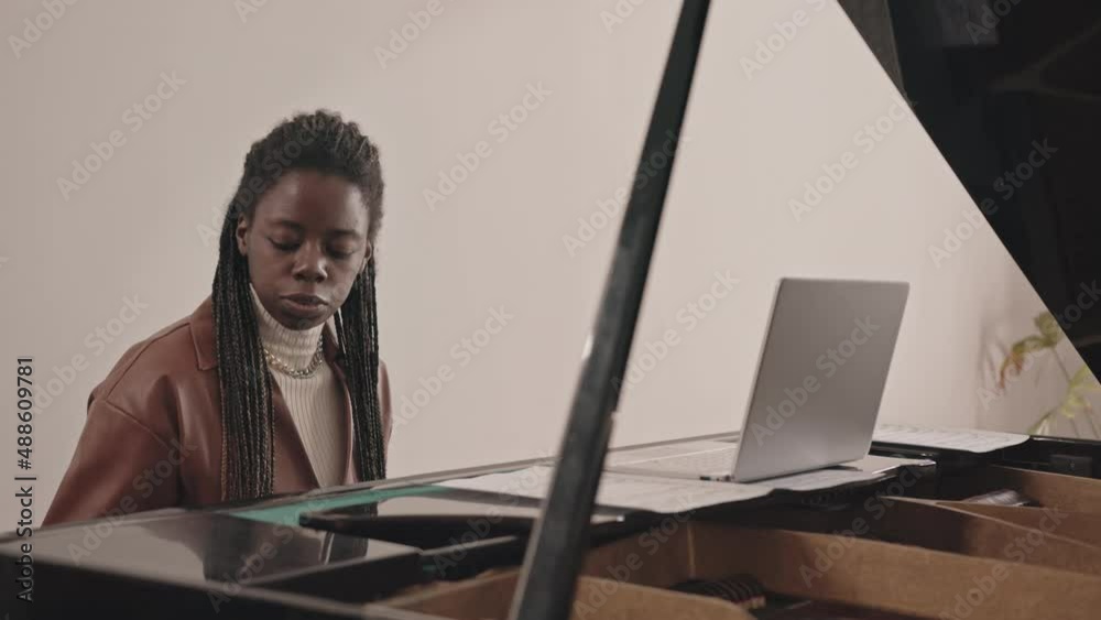 Medium slowmo of young Black woman having online video music lesson on laptop sitting by grand piano indoors