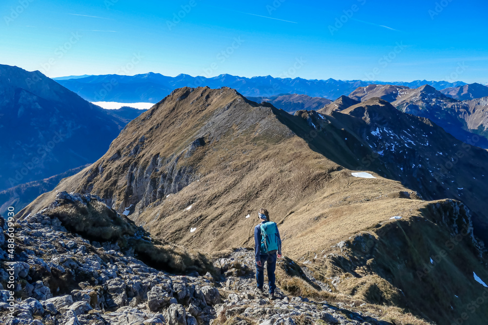 Woman with a backpack walking on a panoramic hiking trail near mount Eisenerzer Reichenstein in Styria, Austria, Europe. Mountain ridges of the Ennstal Alps are bare. Path in sunny autumn. Spring vibe
