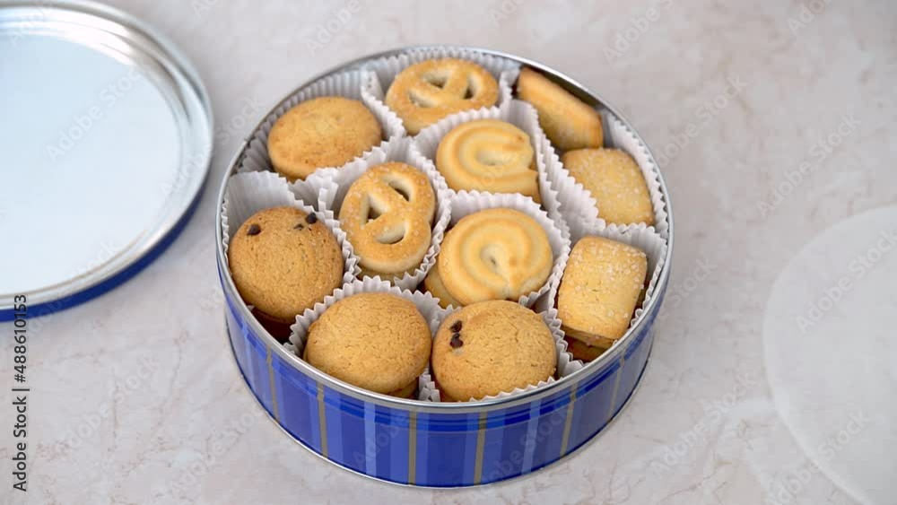 Caucasian woman hand removing parchment paper from full tin of danish butter cookies. Crispy shortbread biscuits in an open container on the table. Sweet breakfast. Top view.