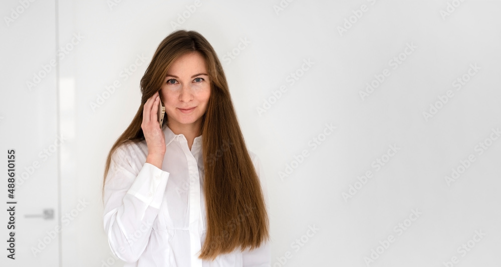 Caucasian woman with long brown hair smiling, speaking by phone,having call looking at camera on white background at home or office.Banner,copy space Caucasian woman with long brown hair smiling, speaking by phone,having call looking at camera on white background at home or office.Banner,copy space