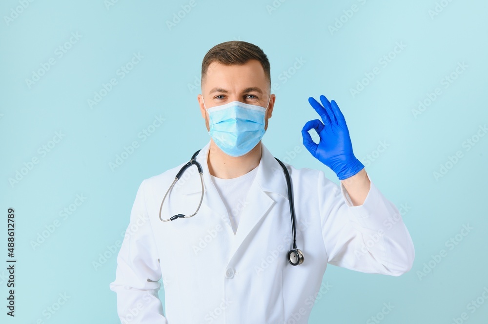 Young male doctor wearing medical mask, and stethoscope round his neck, isolated on blue background