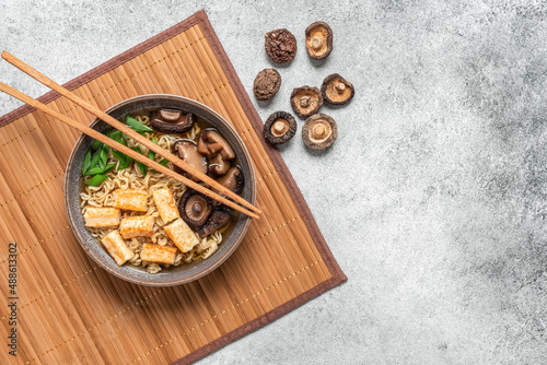 Asian vegan ramen soup with noodles, roasted tofu cheese and shiitake mushrooms in a bowl on a bamboo mat. Gray concrete grunge background. Top view.
