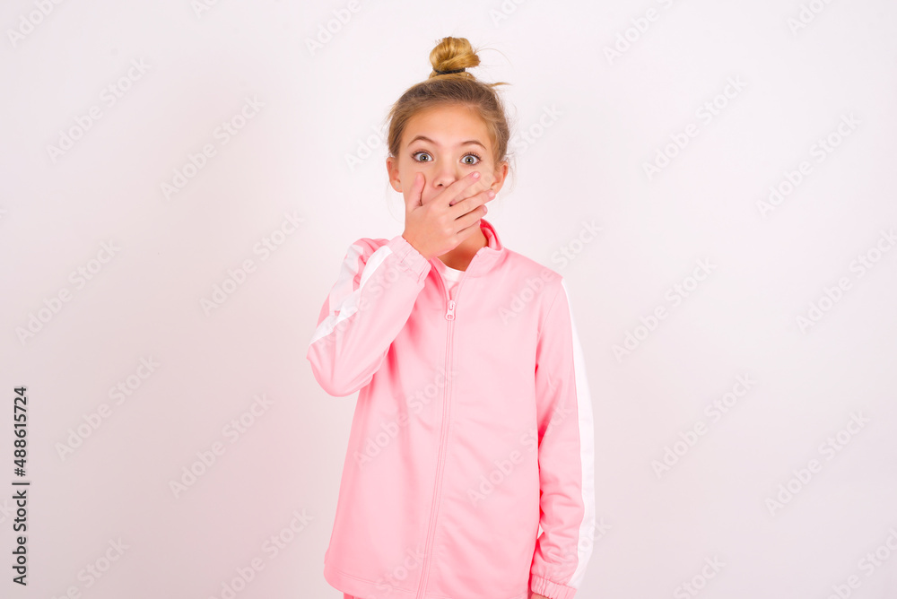 Emotional caucasian little kid girl with bun hairstyle wearing pink tracksuit over white background gasps from astonishment, covers opened mouth with palm, looks shocked at camera. Reaction concept