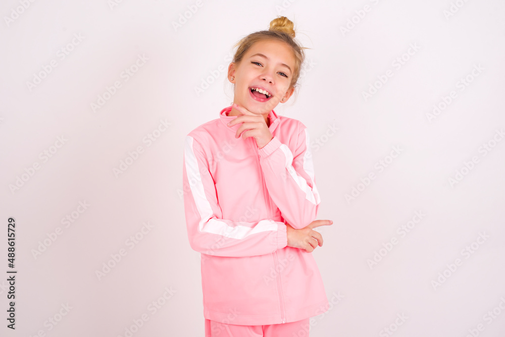 Optimistic caucasian little kid girl with bun hairstyle wearing pink tracksuit over white wall keeps hands partly crossed and hand under chin, looks at camera with pleasure. Happy emotions concept. Optimistic caucasian little kid girl with bun hairstyle wearing pink tracksuit over white wall keeps hands partly crossed and hand under chin, looks at camera with pleasure. Happy emotions concept.