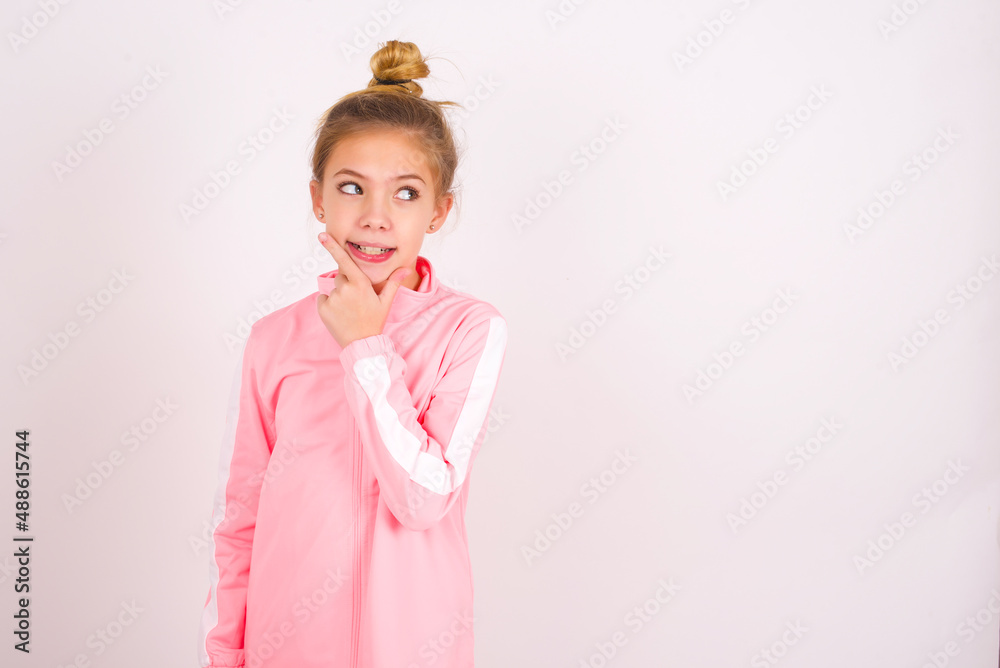 caucasian little kid girl with bun hairstyle wearing pink tracksuit over white background Thinking worried about a question, concerned and nervous with hand on chin. caucasian little kid girl with bun hairstyle wearing pink tracksuit over white background Thinking worried about a question, concerned and nervous with hand on chin.