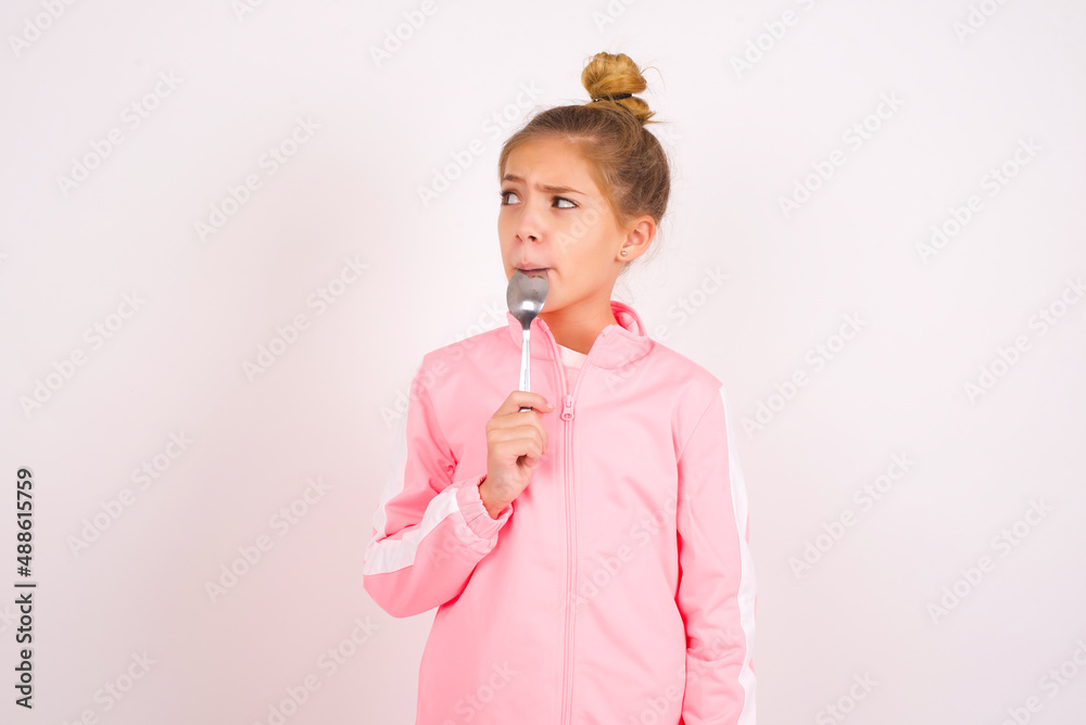 Very hungry caucasian little kid girl with bun hairstyle wearing pink tracksuit over white background holding spoon into mouth dream of tasty meal Very hungry caucasian little kid girl with bun hairstyle wearing pink tracksuit over white background holding spoon into mouth dream of tasty meal