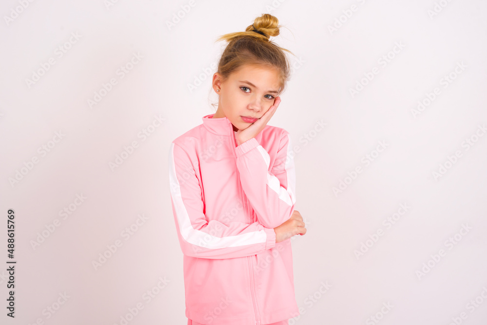 Very bored caucasian little kid girl with bun hairstyle wearing pink tracksuit over white background holding hand on cheek while support it with another crossed hand, looking tired and sick,