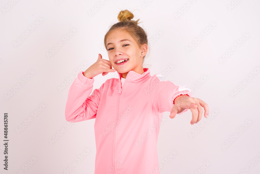 caucasian little kid girl with bun hairstyle wearing pink tracksuit over white background smiling cheerfully and pointing to camera while making a call you later gesture, talking on phone