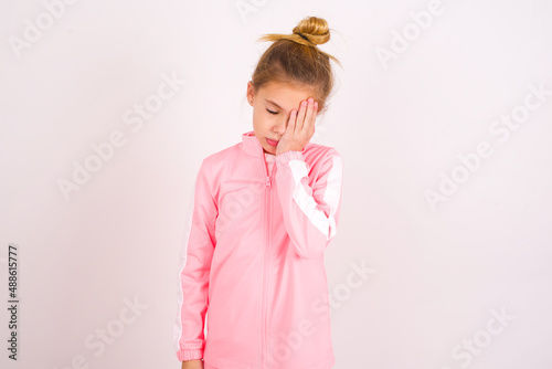 Tired overworked caucasian little kid girl with bun hairstyle wearing pink tracksuit has sleepy expression, gloomy look, covers face with hand, has eyes shut, gasps from tiredness, fatigue after party
