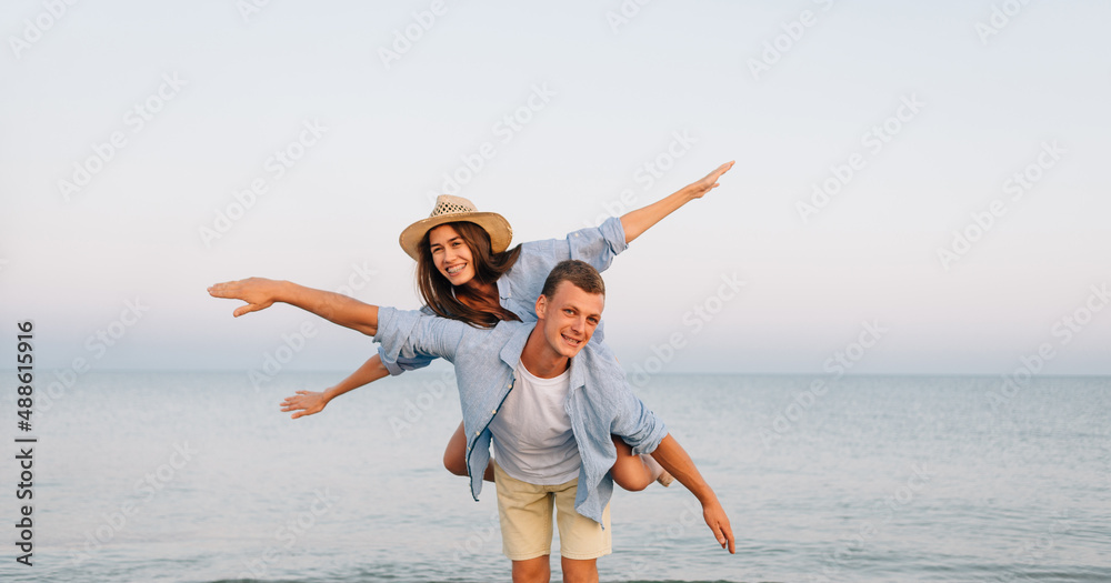Happy man giving piggyback ride to his woman and laughing at tropical beach. Smiling guy in love carrying on back her girlfriend and having fun. Joyful couple enjoying summer at sea