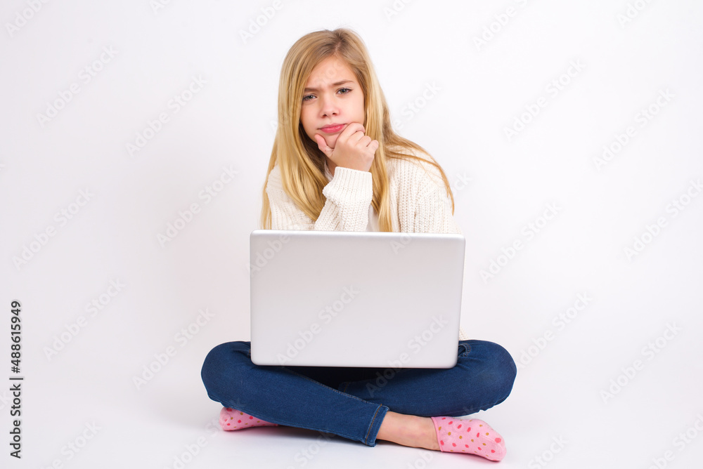 Thoughtful smiling caucasian teen girl sitting with laptop in lotus position on white background keeps hand under chin, looks directly at camera, listens something with interest. Youth concept. Thoughtful smiling caucasian teen girl sitting with laptop in lotus position on white background keeps hand under chin, looks directly at camera, listens something with interest. Youth concept.