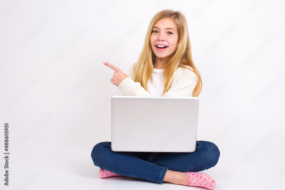 caucasian teen girl sitting with laptop in lotus position on white background pointing away and smiling to you. Look over there! caucasian teen girl sitting with laptop in lotus position on white background pointing away and smiling to you. Look over there!