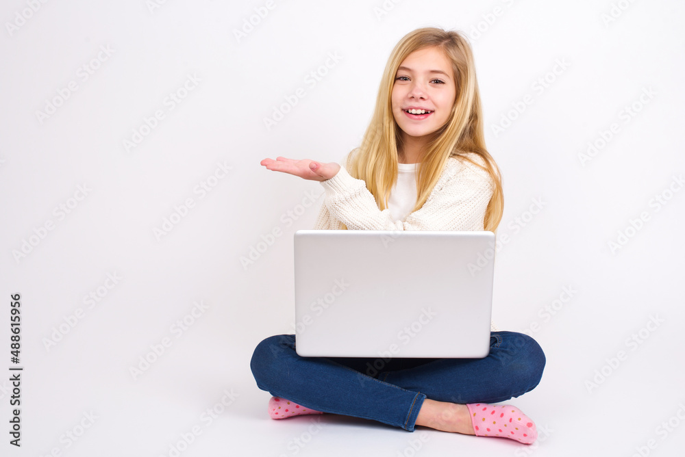 caucasian teen girl sitting with laptop in lotus position on white background says: wow how exciting it is, has amazed expression, shows something on blank space with palm. Advertisement concept. caucasian teen girl sitting with laptop in lotus position on white background says: wow how exciting it is, has amazed expression, shows something on blank space with palm. Advertisement concept.