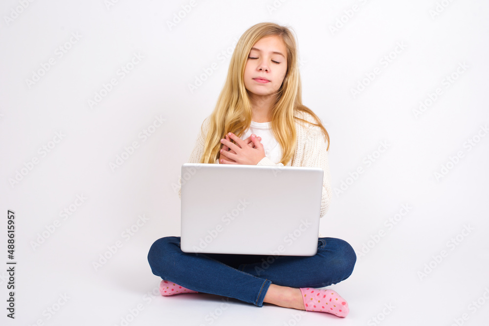 caucasian teen girl sitting with laptop in lotus position on white background closes eyes and keeps hands on chest near heart, expresses sincere emotions, being kind hearted and honest. caucasian teen girl sitting with laptop in lotus position on white background closes eyes and keeps hands on chest near heart, expresses sincere emotions, being kind hearted and honest.