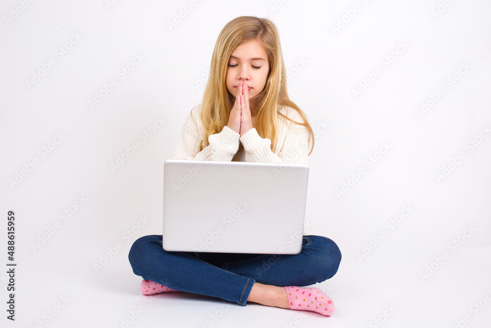 Indoor closeup of caucasian teen girl sitting with laptop in lotus position on white background practicing yoga and meditation, holding palms together in namaste, looking calm, relaxed and peaceful. Indoor closeup of caucasian teen girl sitting with laptop in lotus position on white background practicing yoga and meditation, holding palms together in namaste, looking calm, relaxed and peaceful.