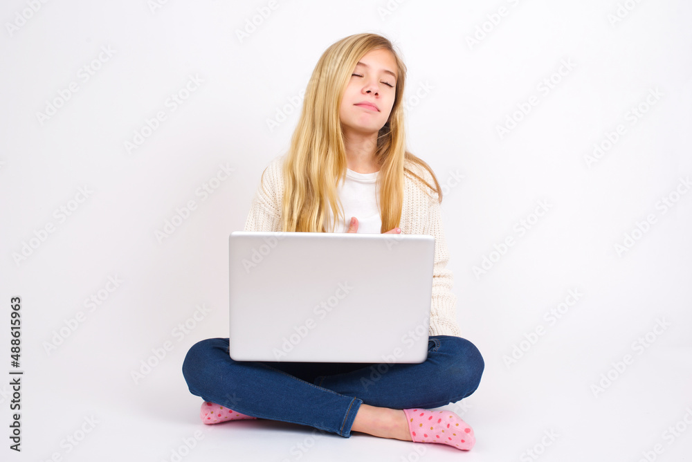 Satisfied smiling caucasian teen girl sitting with laptop in lotus position on white background , keeps hands on belly, being in good mood after eating delicious supper, demonstrates she is full. Satisfied smiling caucasian teen girl sitting with laptop in lotus position on white background , keeps hands on belly, being in good mood after eating delicious supper, demonstrates she is full.
