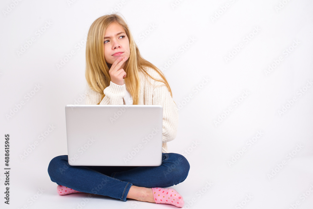 Shot of contemplative thoughtful caucasian teen girl sitting with laptop in lotus position on white background keeps hand under chin, looks thoughtfully upwards. Shot of contemplative thoughtful caucasian teen girl sitting with laptop in lotus position on white background keeps hand under chin, looks thoughtfully upwards.