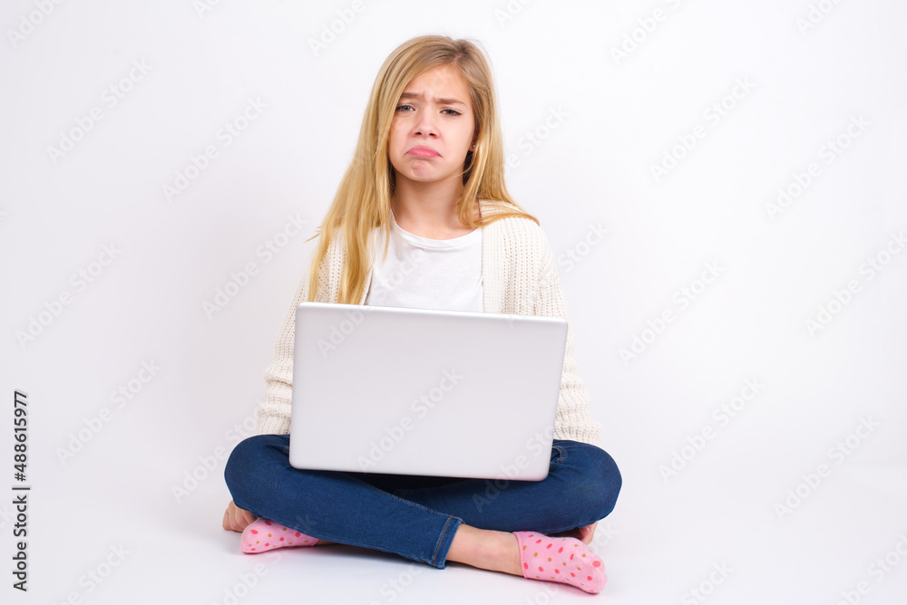 caucasian teen girl sitting with laptop in lotus position on white wall crying desperate and depressed with tears on his eyes suffering pain and depression. Sad facial expression and emotion concept. caucasian teen girl sitting with laptop in lotus position on white wall crying desperate and depressed with tears on his eyes suffering pain and depression. Sad facial expression and emotion concept.