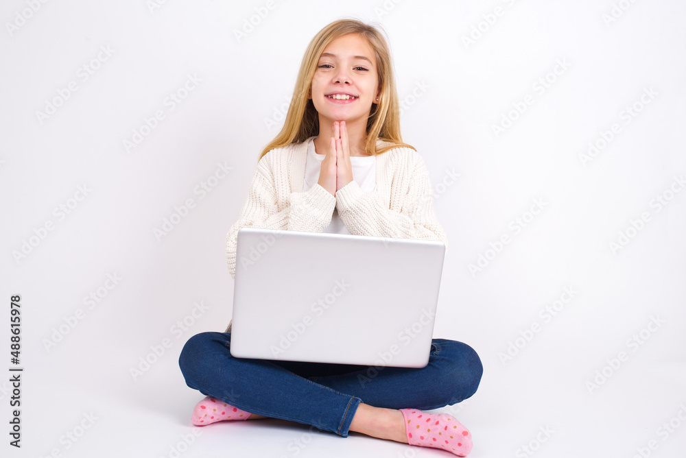 caucasian teen girl sitting with laptop in lotus position on white background keeps palms together, has pleased expression. Glad attractive male makes request, pleads for mercy. Hopeful young adult. caucasian teen girl sitting with laptop in lotus position on white background keeps palms together, has pleased expression. Glad attractive male makes request, pleads for mercy. Hopeful young adult.