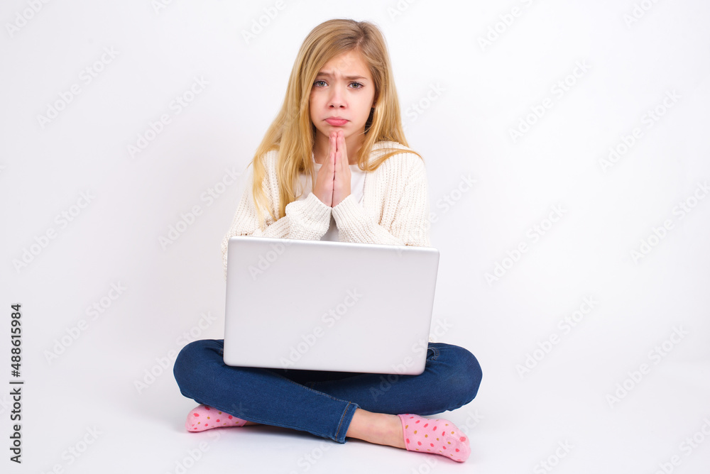 caucasian teen girl sitting with laptop in lotus position on white background keeps palms pressed together in front of her having regretful look, asking for forgiveness. Forgive me please. caucasian teen girl sitting with laptop in lotus position on white background keeps palms pressed together in front of her having regretful look, asking for forgiveness. Forgive me please.
