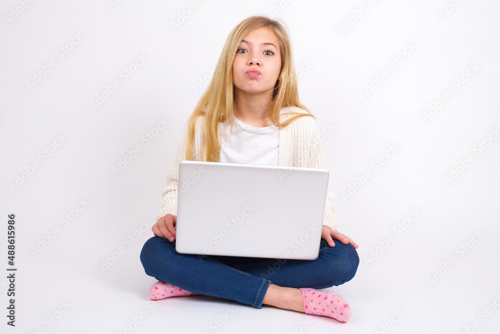 caucasian teen girl sitting with laptop in lotus position on white background , keeps lips as going to kiss someone, has glad expression, grimace face. Standing indoors. Beauty concept. caucasian teen girl sitting with laptop in lotus position on white background , keeps lips as going to kiss someone, has glad expression, grimace face. Standing indoors. Beauty concept.
