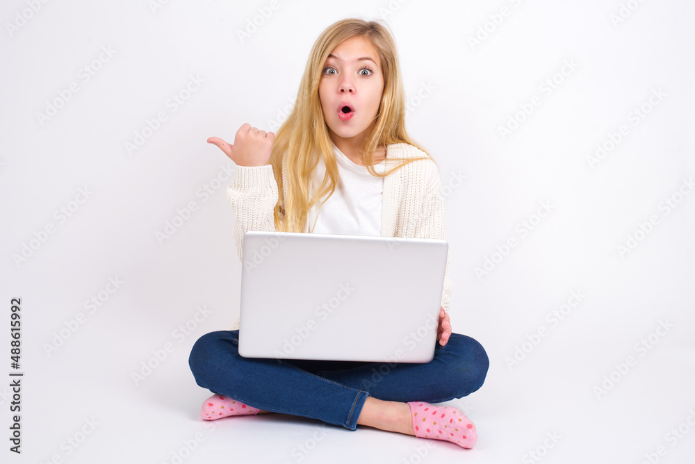 Shocked caucasian teen girl sitting with laptop in lotus position on white background points with thumb away, indicates something. Check this out. Advertisement concept. Shocked caucasian teen girl sitting with laptop in lotus position on white background points with thumb away, indicates something. Check this out. Advertisement concept.