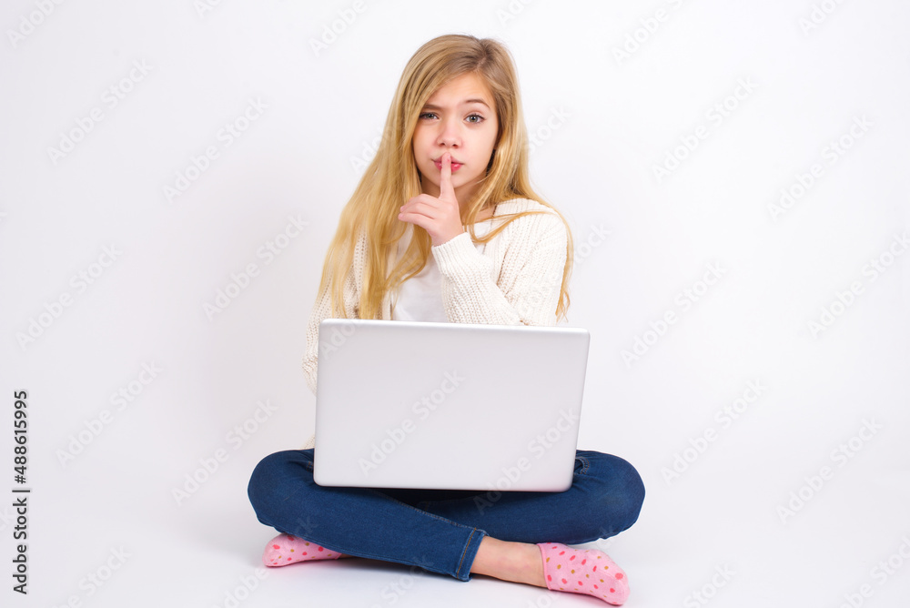 caucasian teen girl sitting with laptop in lotus position on white background makes hush gesture, asks be quiet. Don't tell my secret or not speak too loud, please! caucasian teen girl sitting with laptop in lotus position on white background makes hush gesture, asks be quiet. Don't tell my secret or not speak too loud, please!