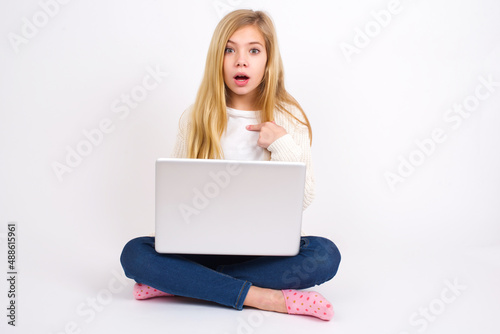 caucasian teen girl sitting with laptop in lotus position on white background being in stupor shocked, has astonished expression pointing at oneself with finger saying: Who me? © Jihan