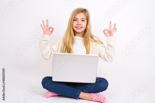 Glad caucasian teen girl sitting with laptop in lotus position on white background shows ok sign with both hands as expresses approval, has cheerful expression, being optimistic. © Jihan
