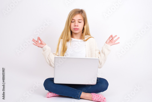 caucasian teen girl sitting with laptop in lotus position on white background doing yoga, keeping eyes closed, holding fingers in mudra gesture. Meditation, religion and spiritual practices.