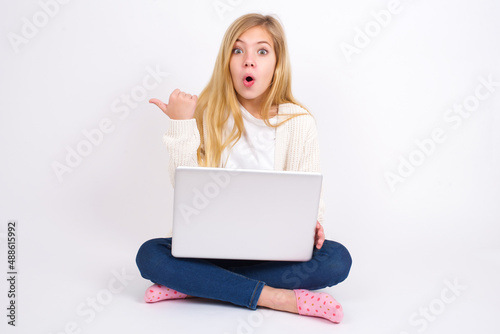 Shocked caucasian teen girl sitting with laptop in lotus position on white background points with thumb away, indicates something. Check this out. Advertisement concept. © Jihan