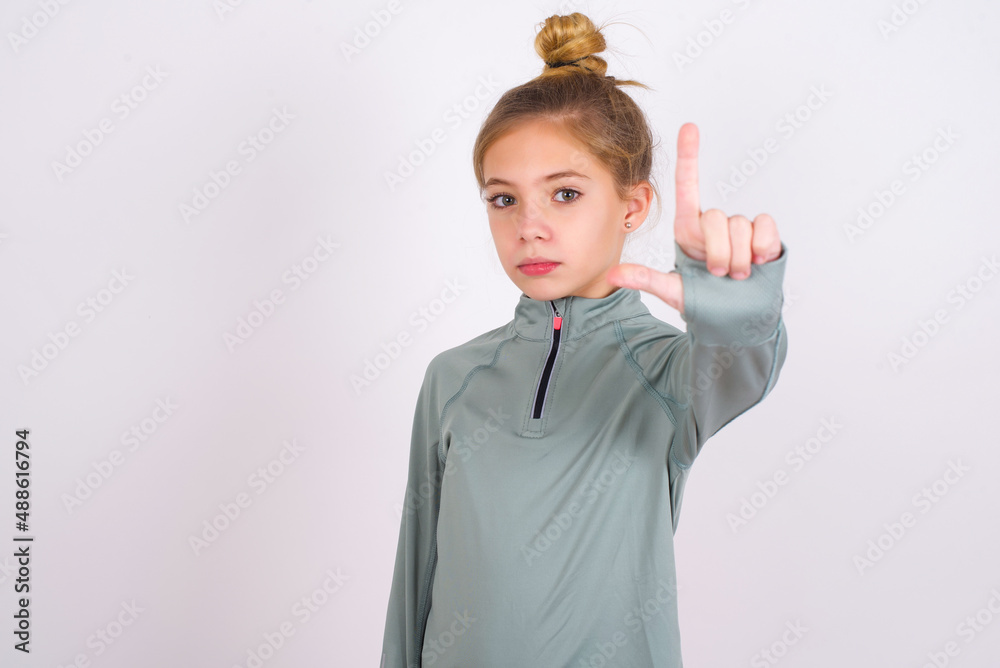little caucasian kid girl with hair bun wearing technical shirt over white background making fun of people with fingers on forehead doing loser gesture mocking and insulting. little caucasian kid girl with hair bun wearing technical shirt over white background making fun of people with fingers on forehead doing loser gesture mocking and insulting.
